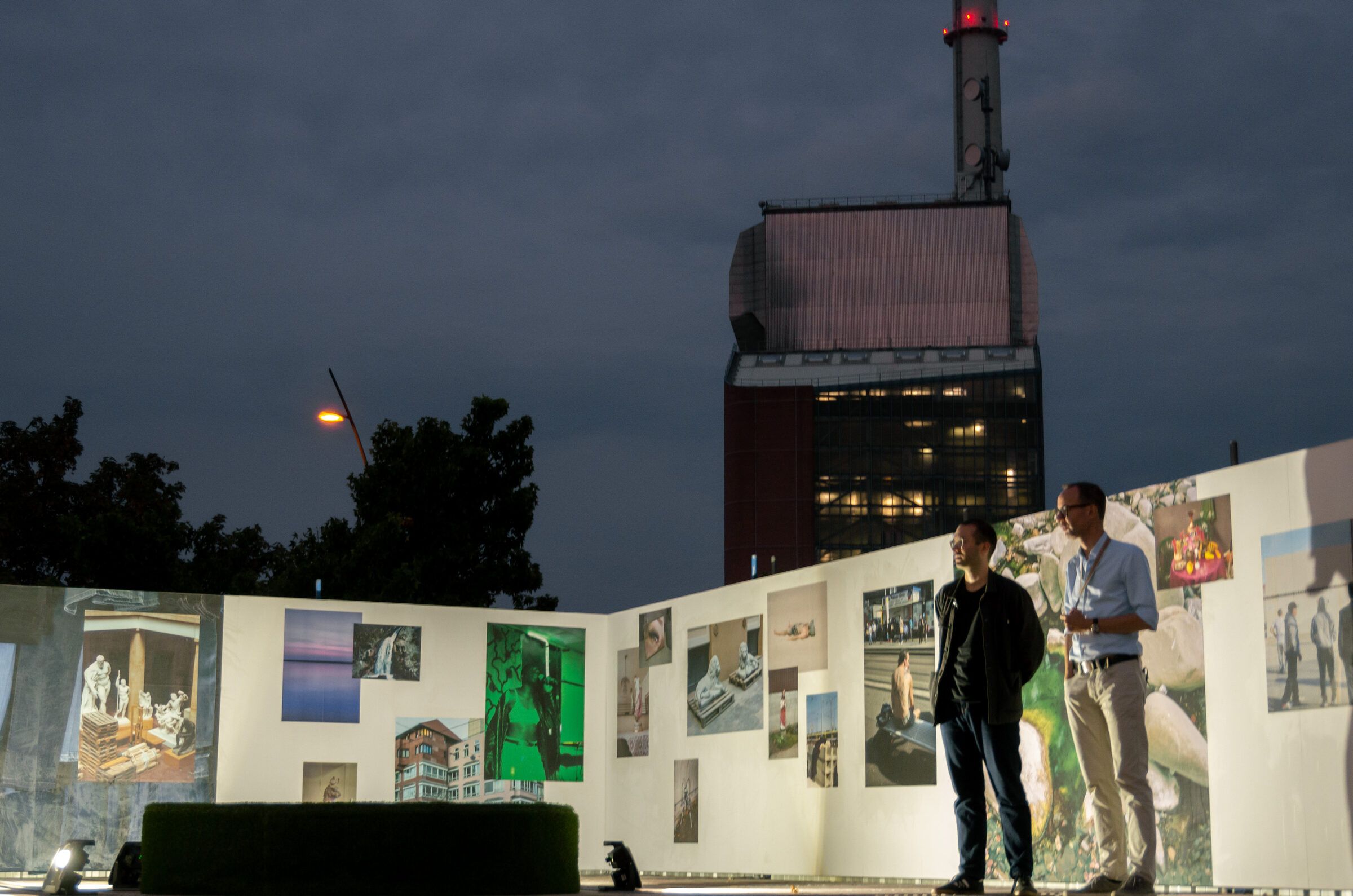 In der Abenddämmerung stehen zwei Personen vor einer beleuchteten Ausstellungswand, dahinter das Kraftwerk.
