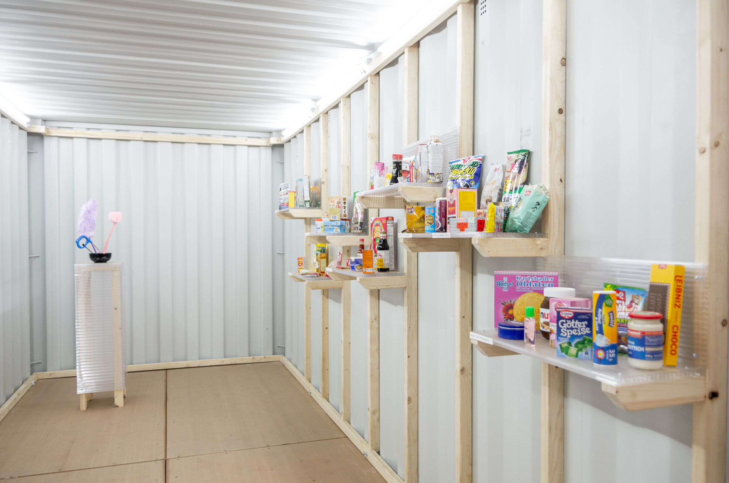 Ein heller Containerraum mit Regalbrettern, auf denen farbige Objekte ausgestellt sind.