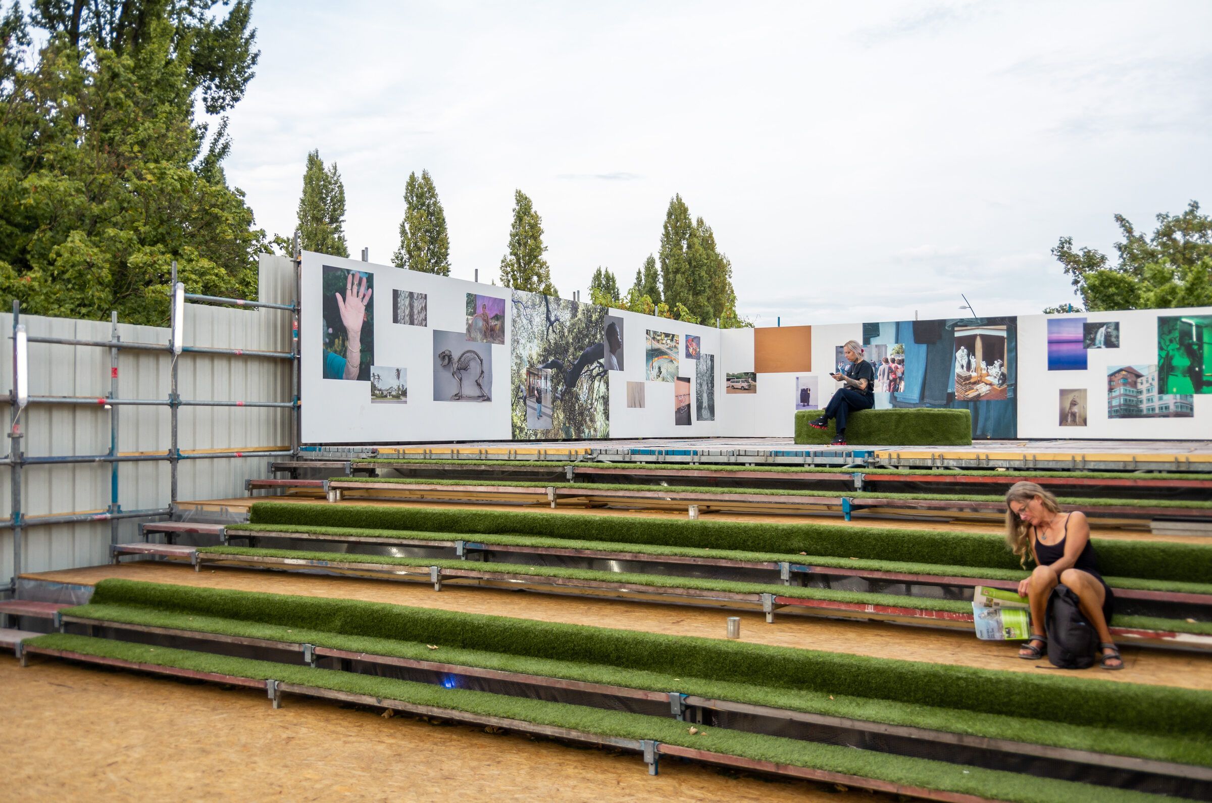 Eine grün bespannte Stufentribüne vor einer langen Ausstellungswand mit großformatigen Bildern unter blauem Himmel.