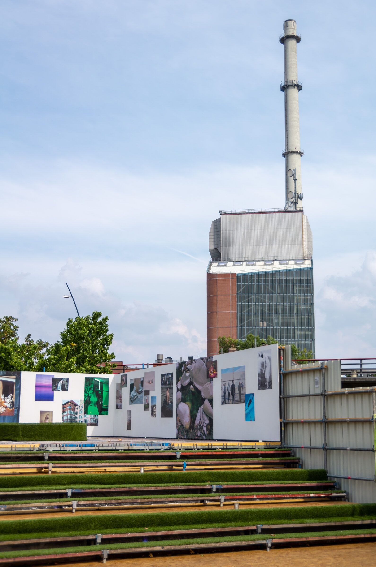 Hochformat mit grüner Sitztribüne, Ausstellungswand und dem Kraftwerksschornstein im Hintergrund.