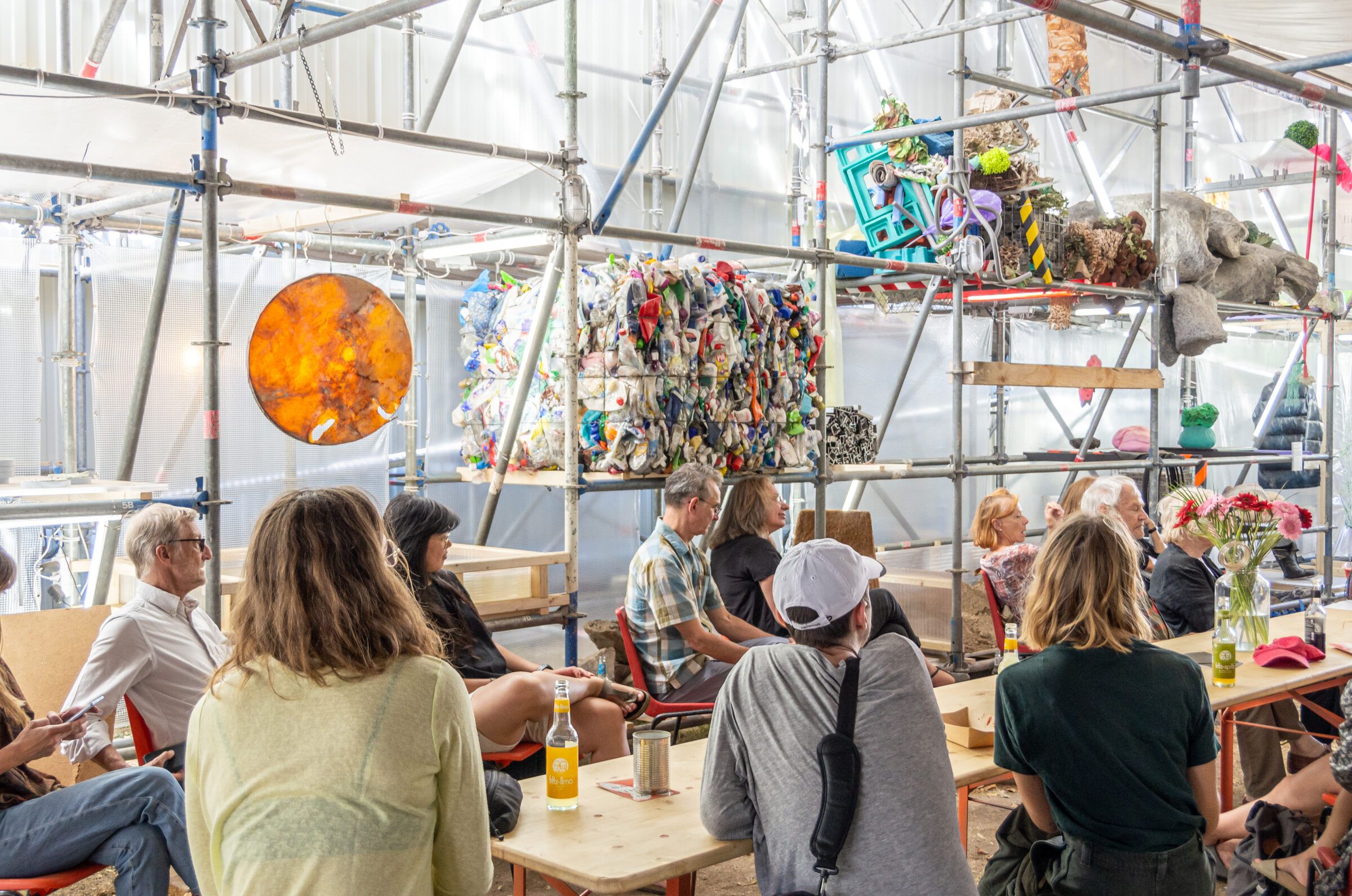 Besuchende sitzen an der langen Tafel unter dem Gerüstdach, über ihnen hängen Objekte der Materialbibliothek.