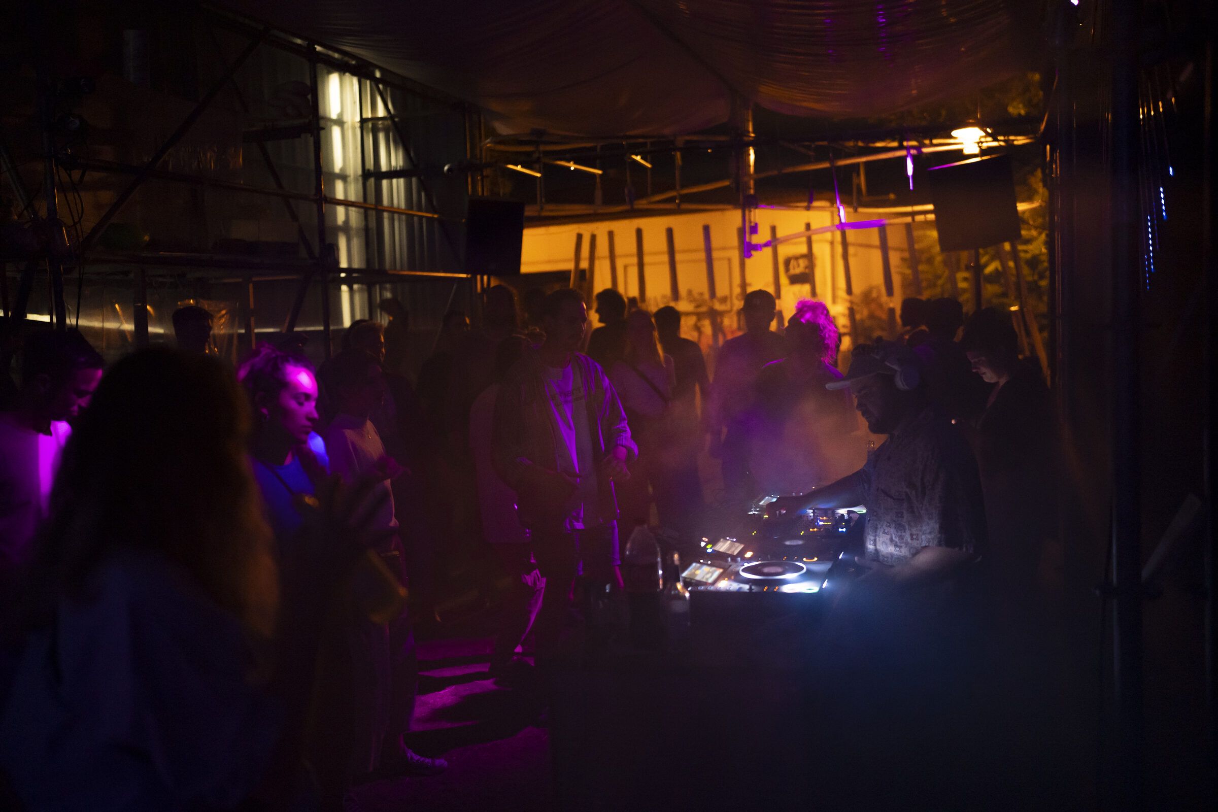 Blick in die nächtliche Gerüsthalle mit violettem Licht, Silhouetten von Gästen und einem DJ-Pult.