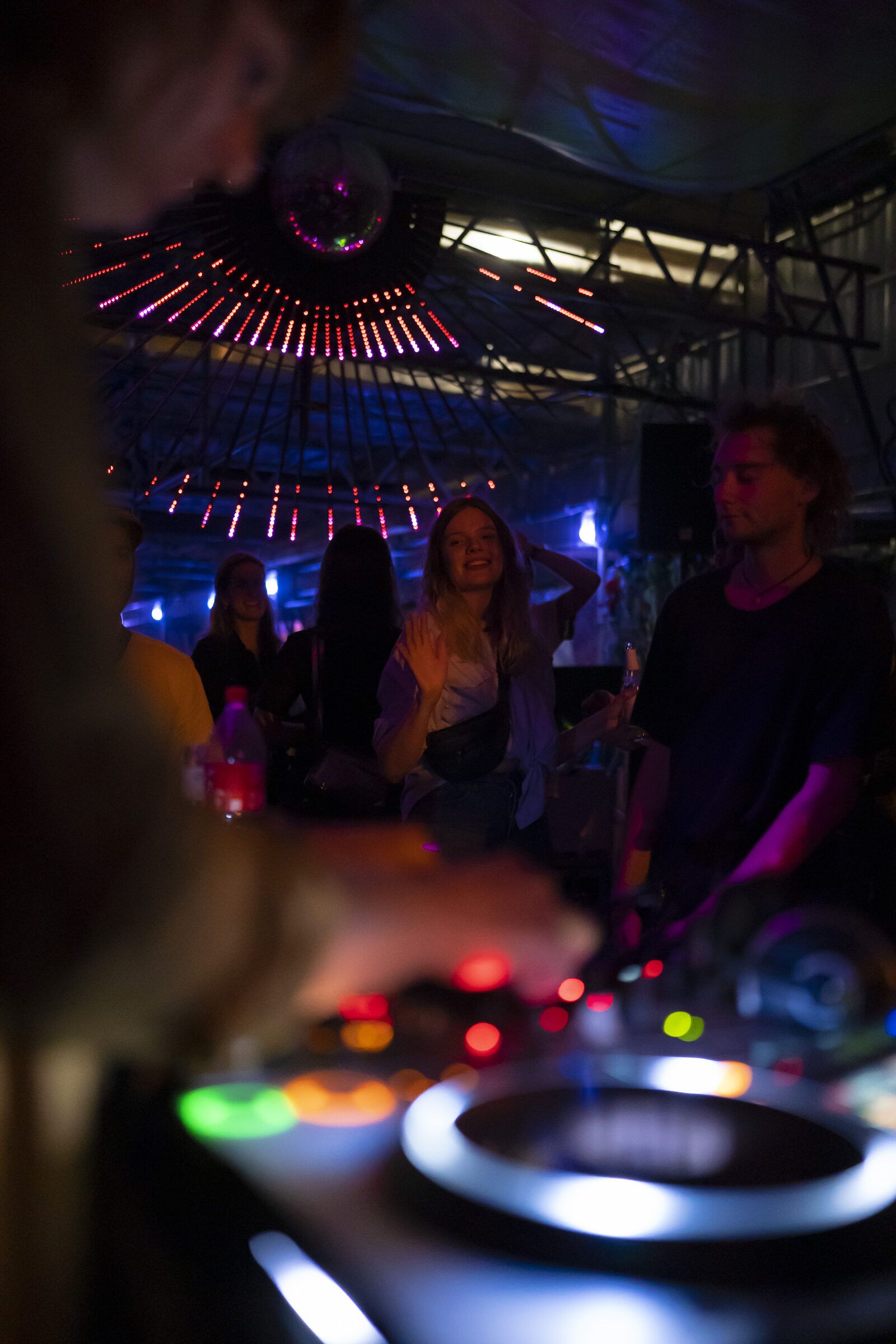 Nahaufnahme eines DJ-Pults mit leuchtenden Reglern, im Hintergrund Gäste im Licht.