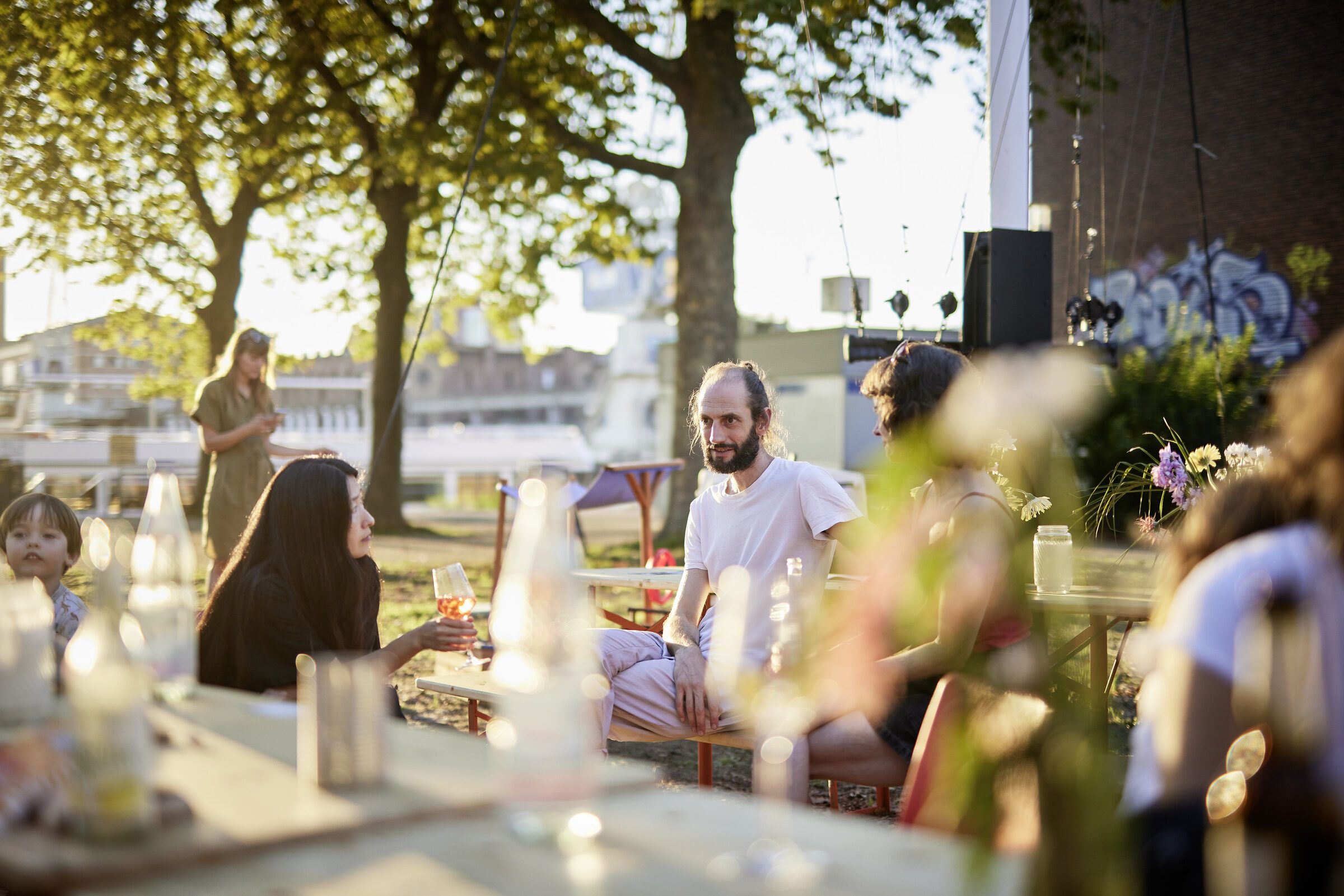 Menschen sitzen an Tischen unter Bäumen am Hafenufer, im Vordergrund Gläser und Blumen.