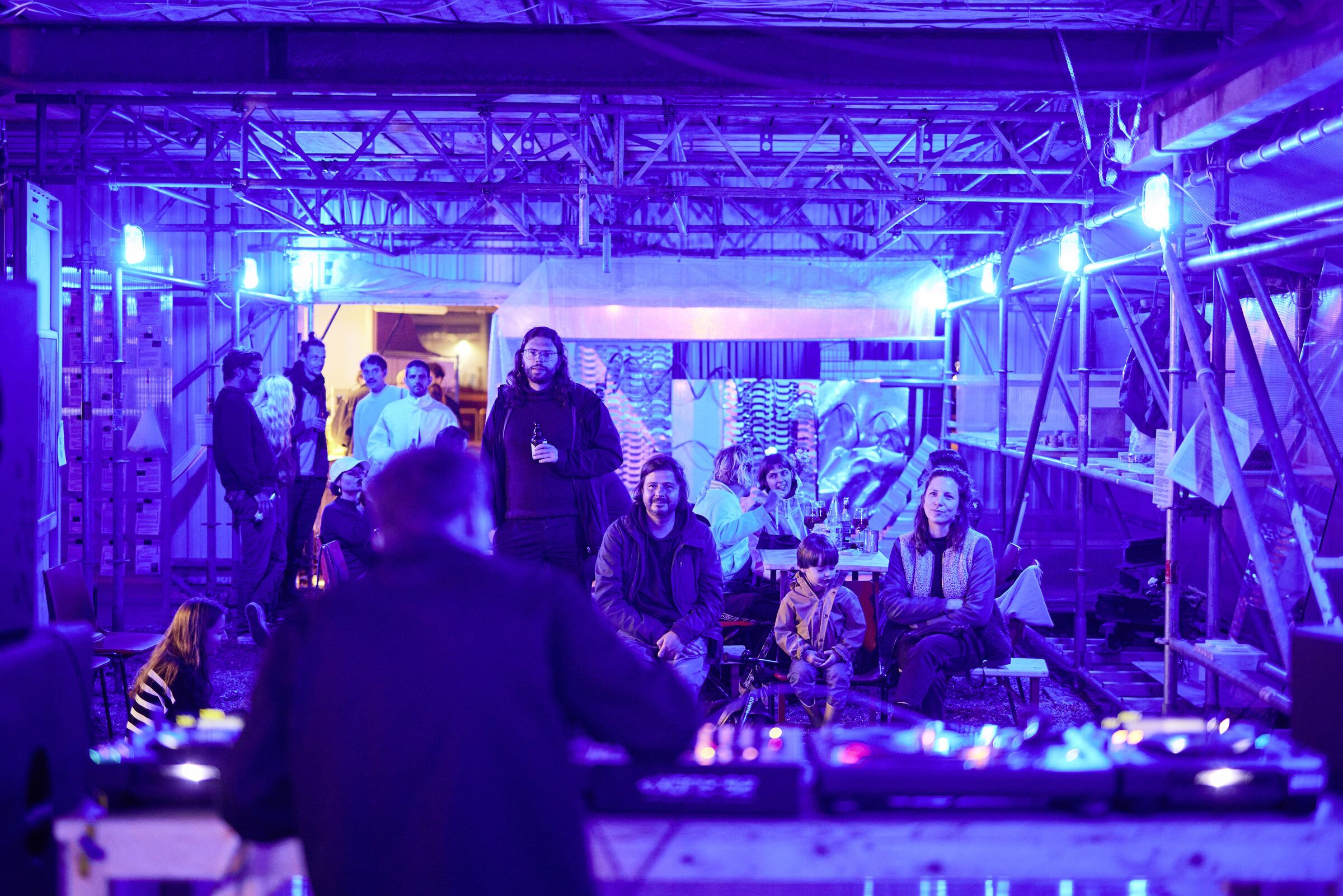 Gäste stehen und sitzen um das blau beleuchtete DJ-Pult in der Halle.