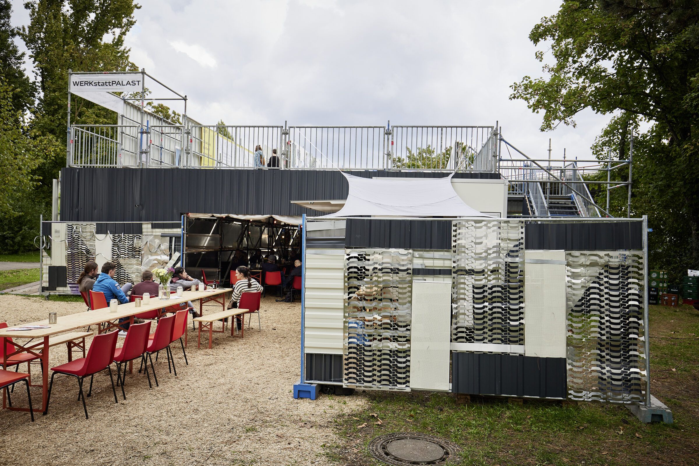 Außenansicht mit bedruckter Containerfassade, dahinter der Gerüstbau mit Dachterrasse.