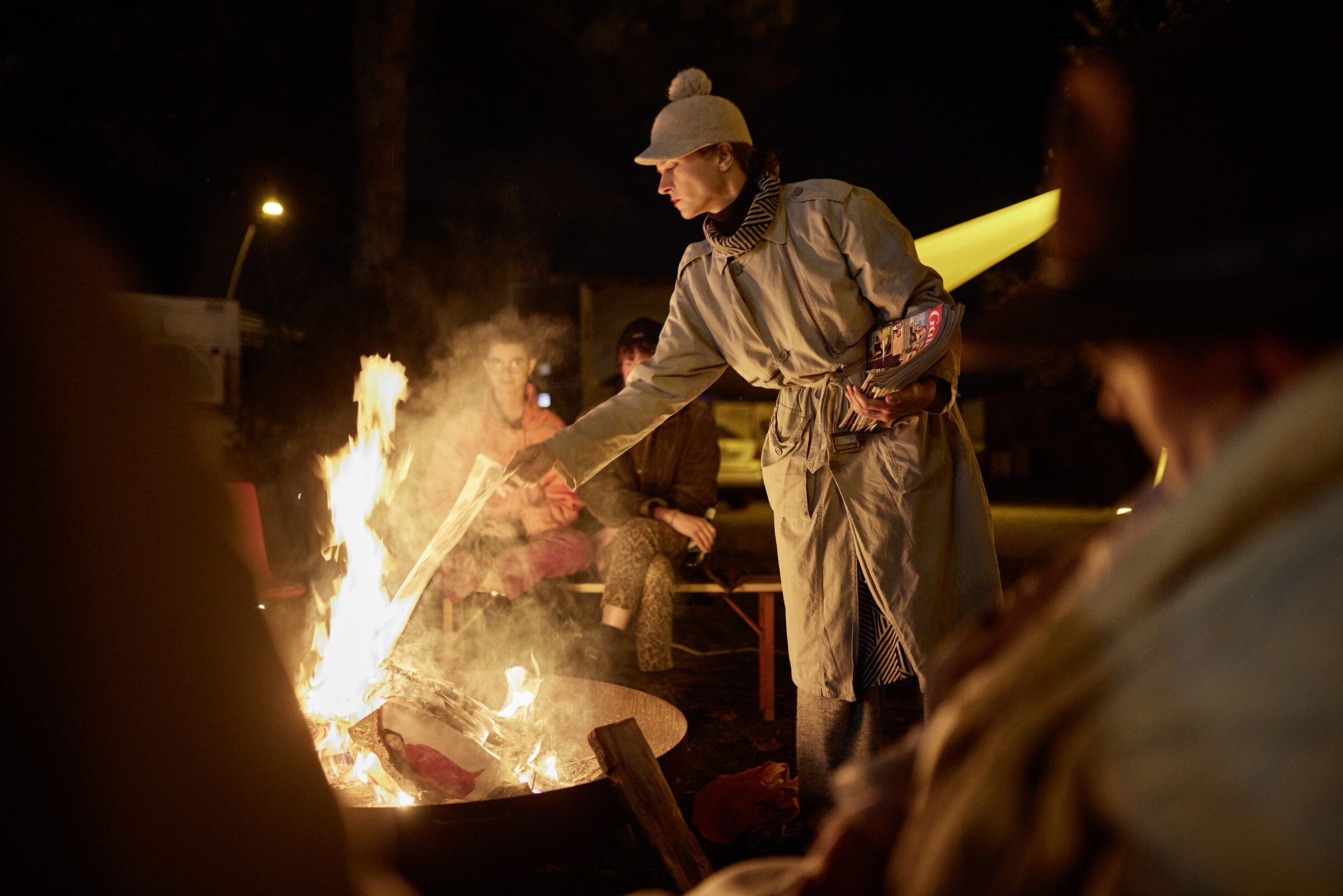 Ein Performer in hellem Anzug steht an einer Feuerschale.