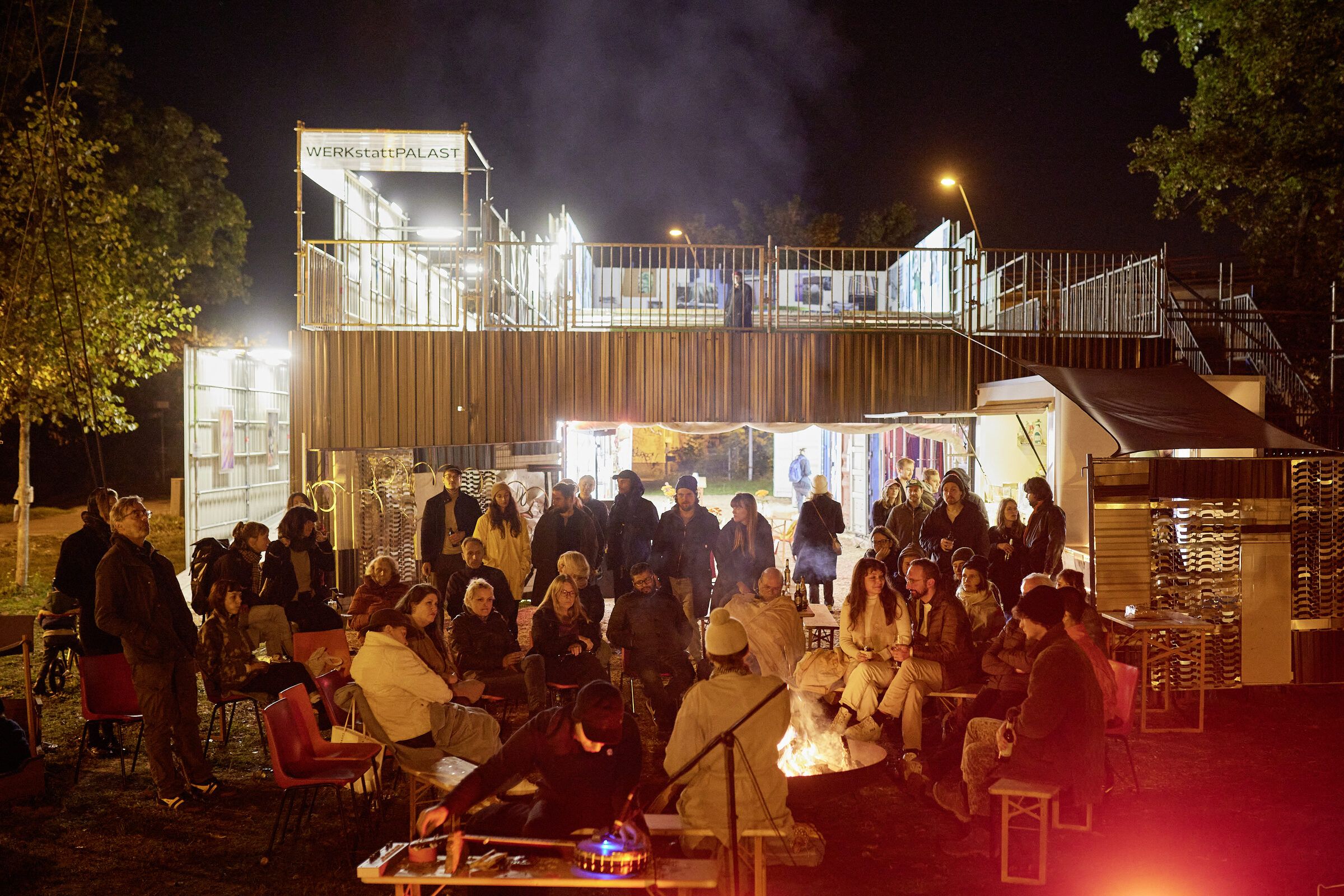 Publikum sitzt nachts um ein Feuer vor dem beleuchteten Gerüstbau.