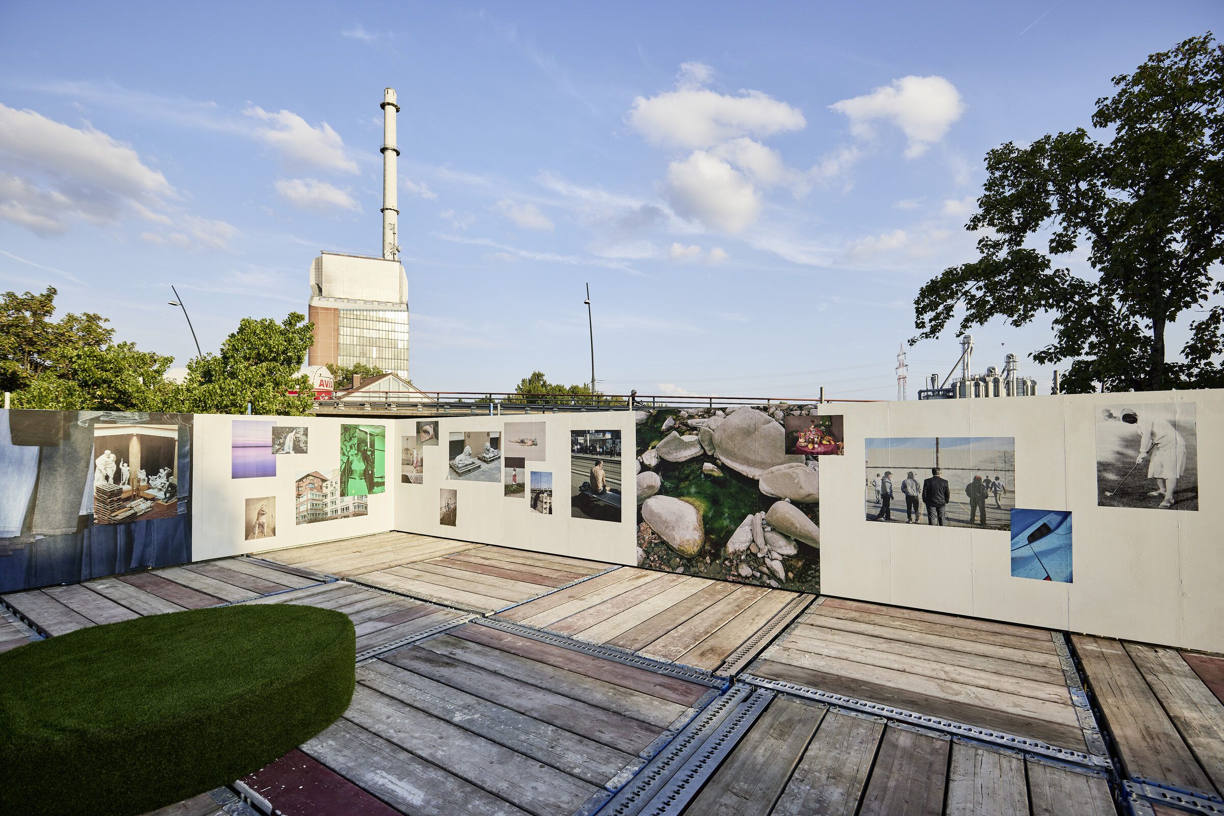 Fotowände der Ausstellung auf der Dachterrasse unter blauem Himmel, dahinter ein Industrieschornstein.