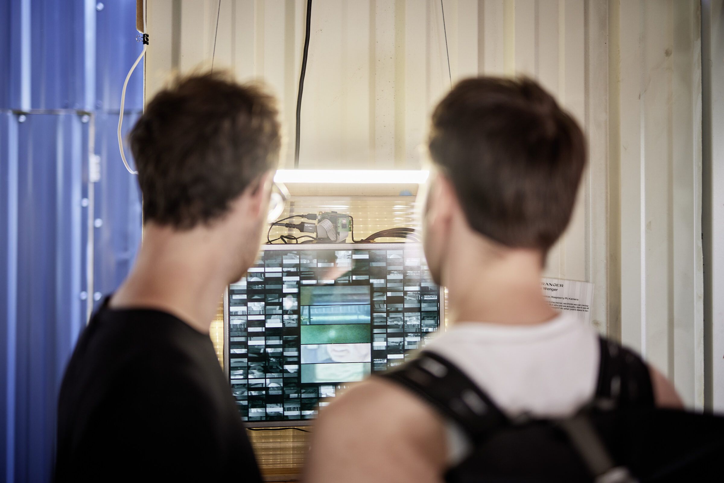 Zwei junge Männer betrachten von hinten eine Bildschirminstallation mit Kamera.