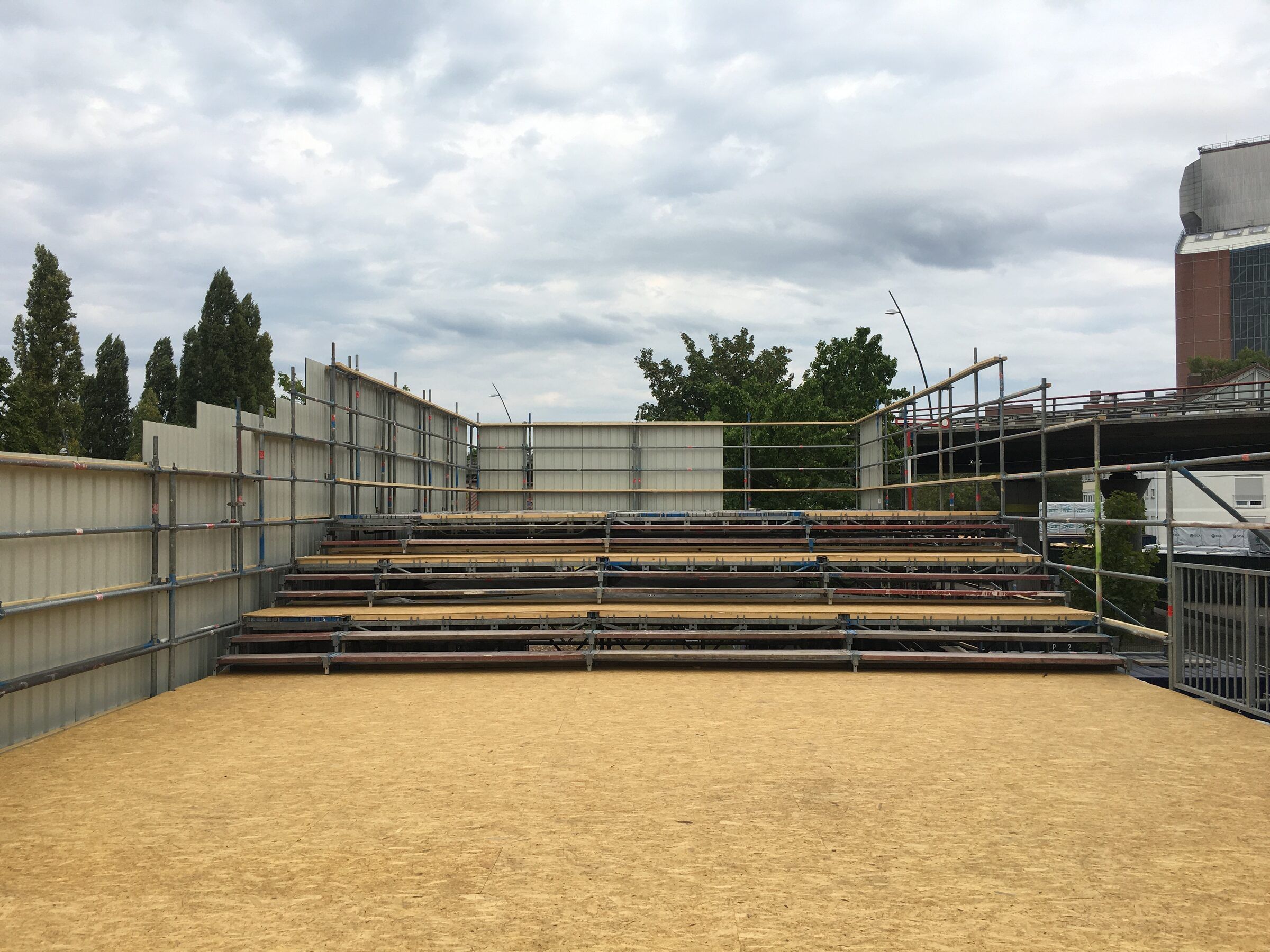Die fertige Tribüne auf dem Dach, noch leer, mit sandfarbenem Boden und grauem Himmel.