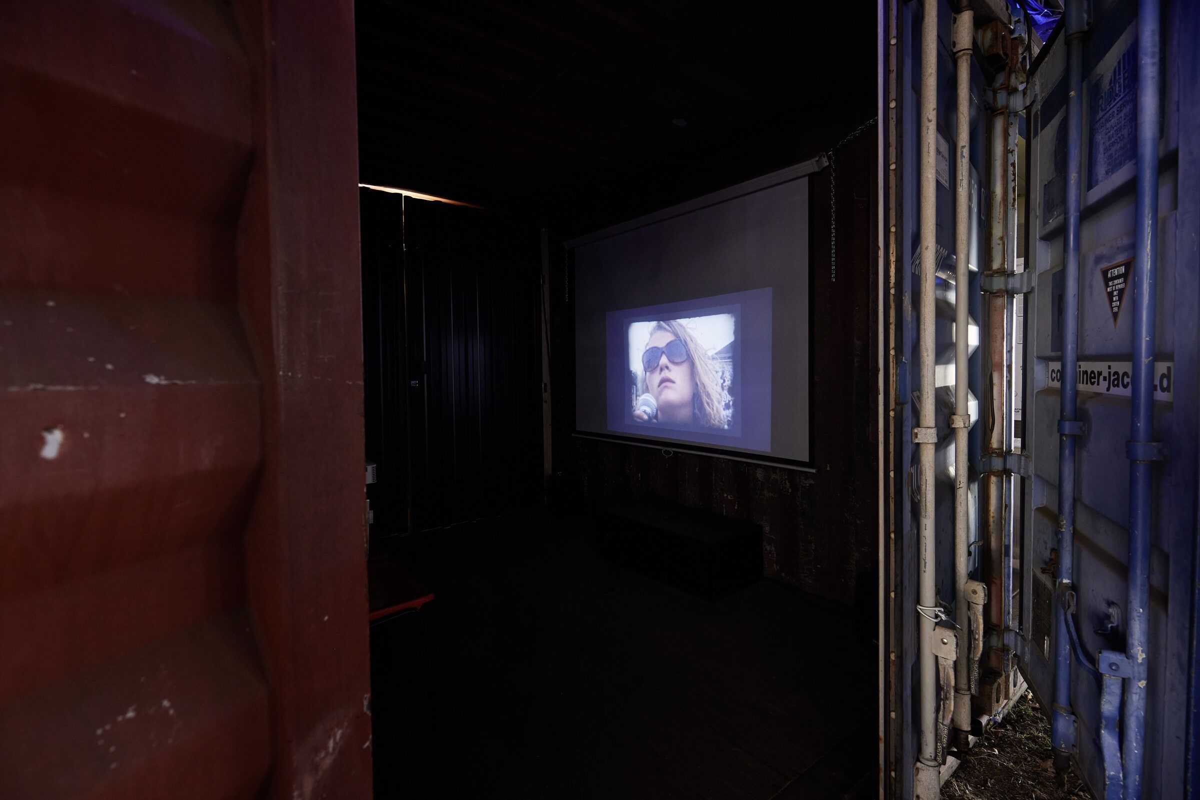 Blick in einen dunklen Container, in dem ein Video auf eine Leinwand projiziert wird.