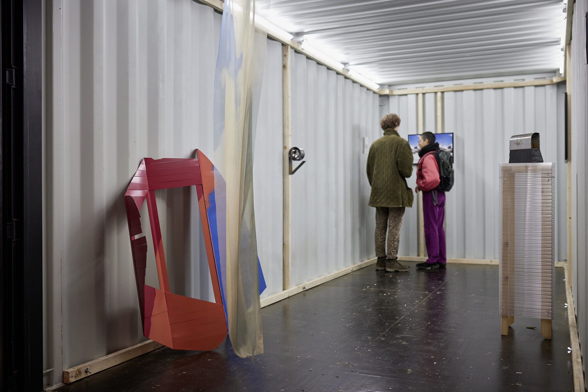 Zwei Besucher:innen im hellen Container, vorne eine rote, gefaltete Skulptur.