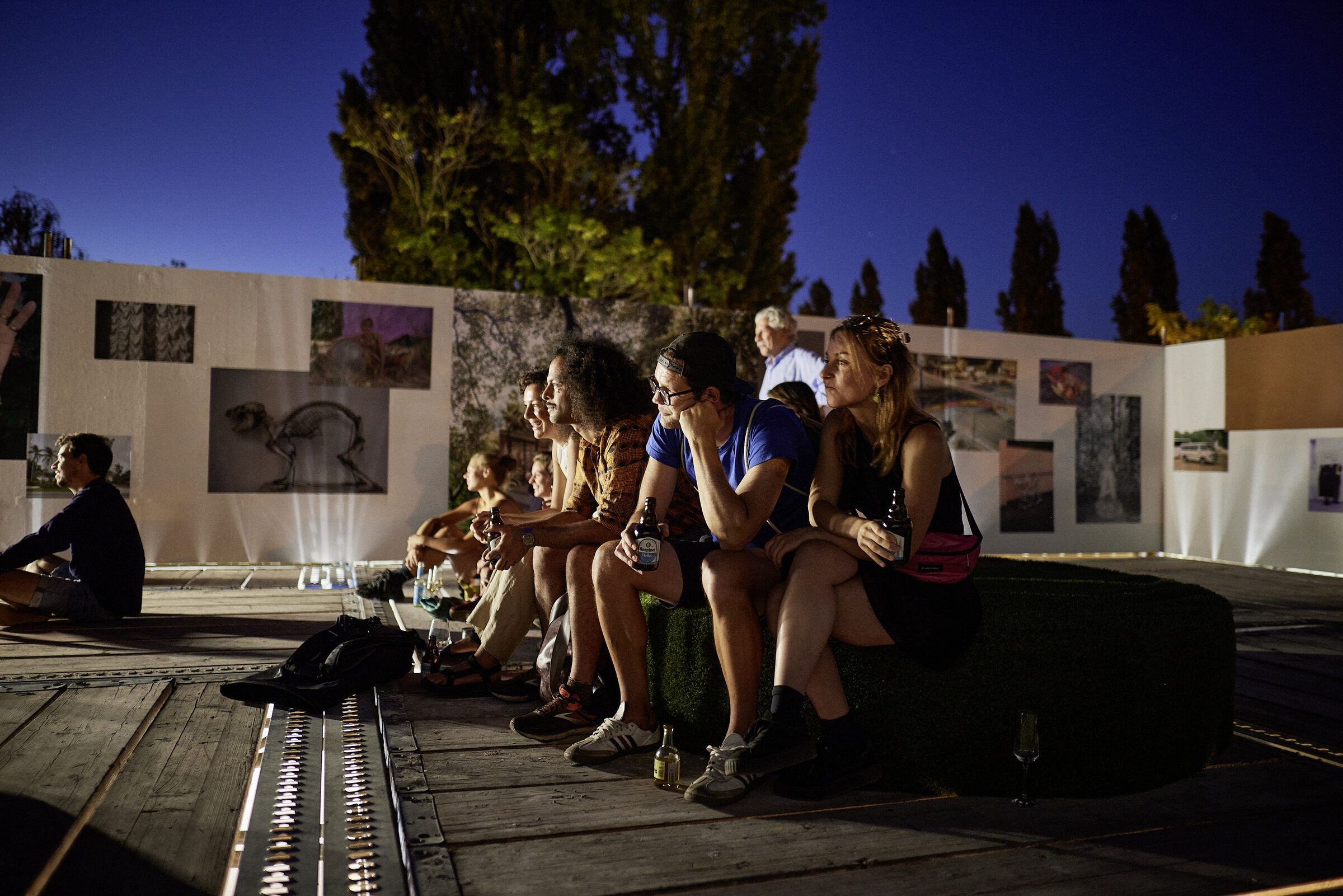 Menschen sitzen nachts auf der Dachterrasse vor den Fotowänden, im Hintergrund dunkelblauer Himmel und Pappeln.