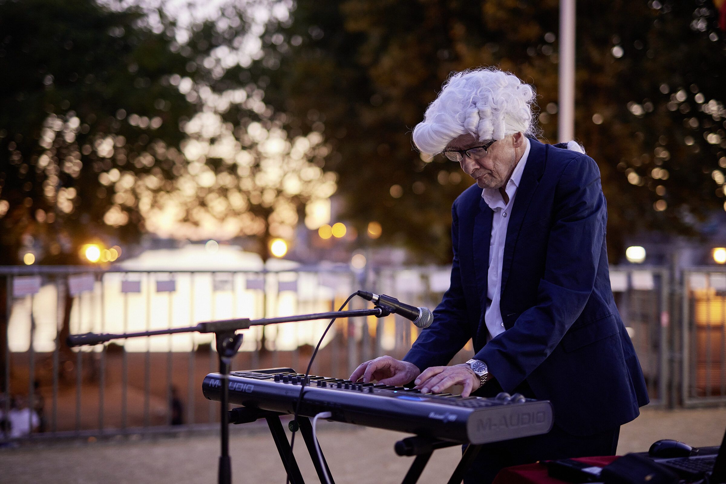 Ein Musiker mit weißem Haar spielt am Wasser Keyboard, im Hintergrund leuchtet die Abendsonne durch die Bäume.