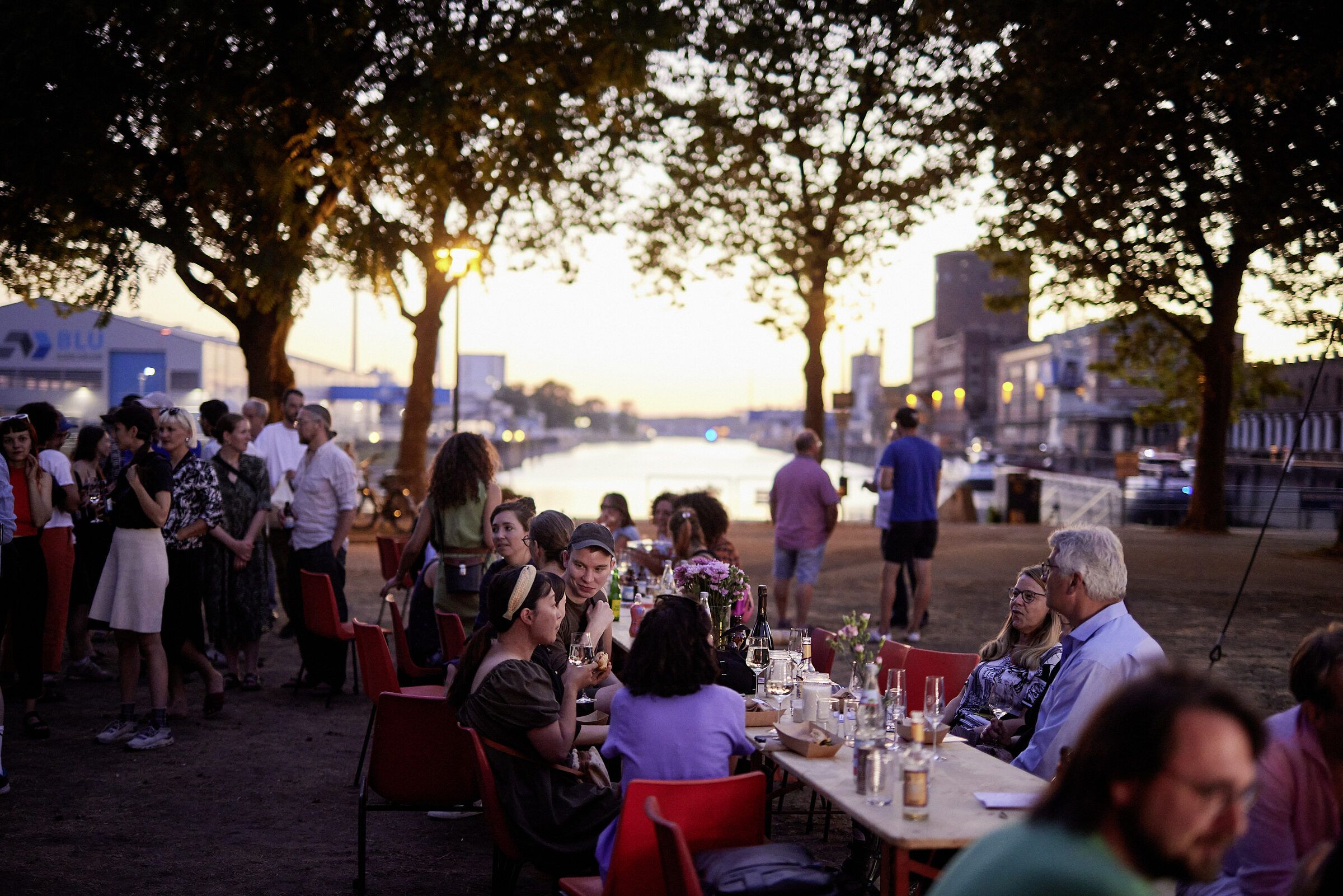 Gäste an der langen Tafel am Wasser, die tiefstehende Sonne leuchtet durch die Bäume.