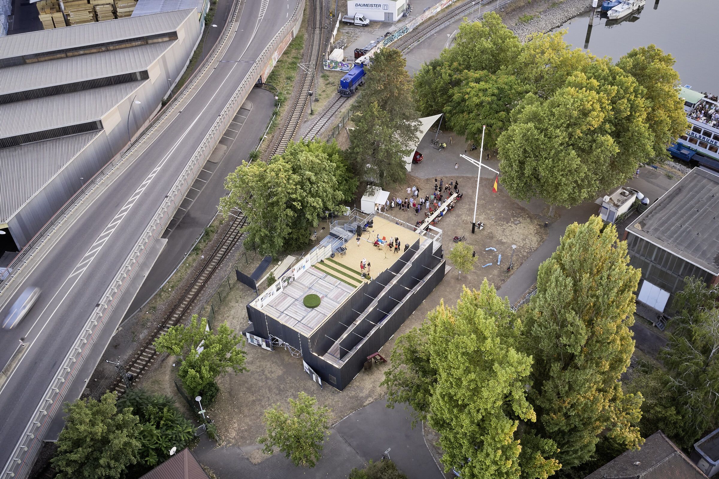 Draufsicht von oben auf das Gebäude mit Dachterrasse, umgeben von Bäumen, Gleisen und einer Straße.
