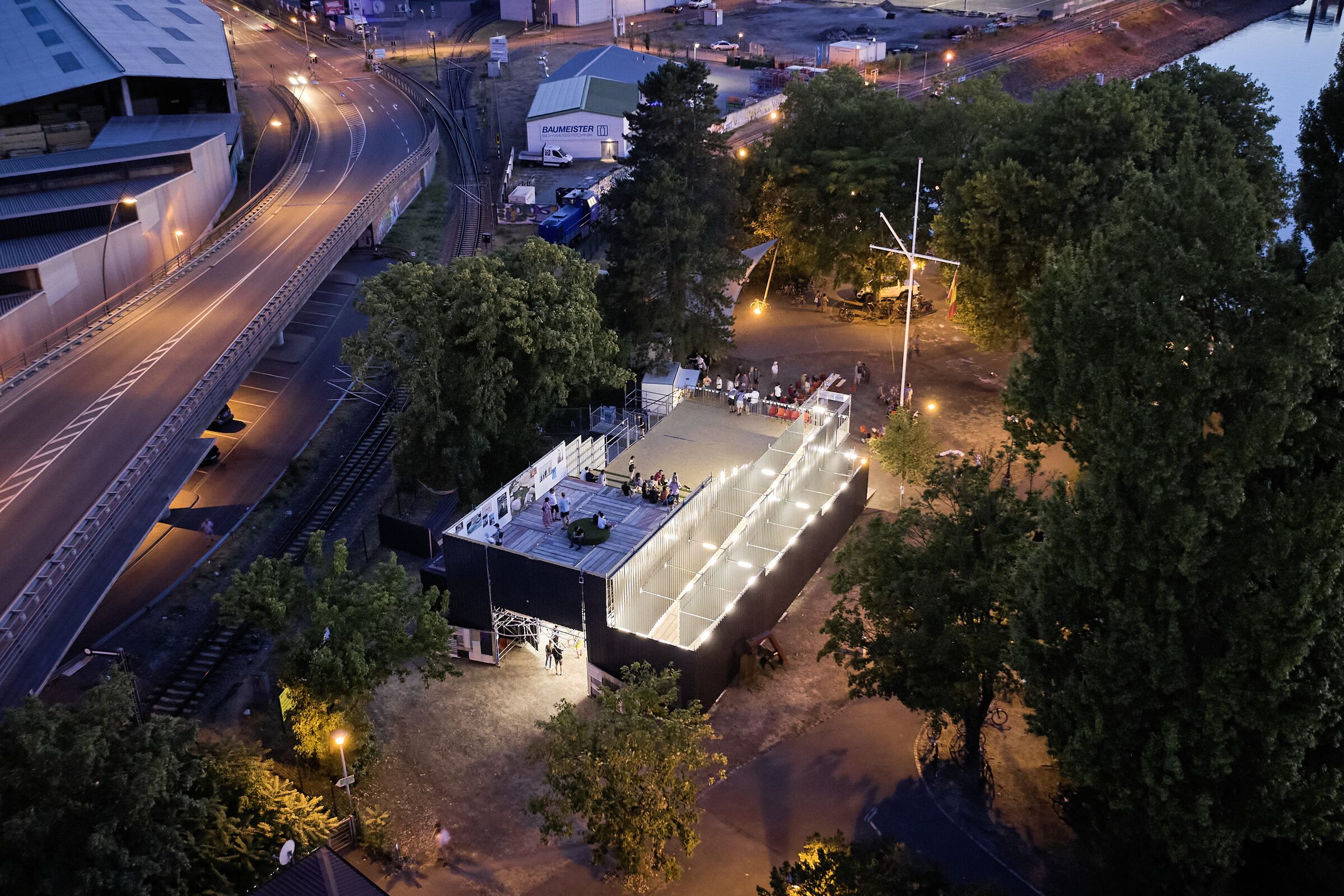Drohnenaufnahme bei Nacht: Das hell beleuchtete Gerüstgebäude steht zwischen Bäumen, daneben eine Straße mit Lichtspuren.