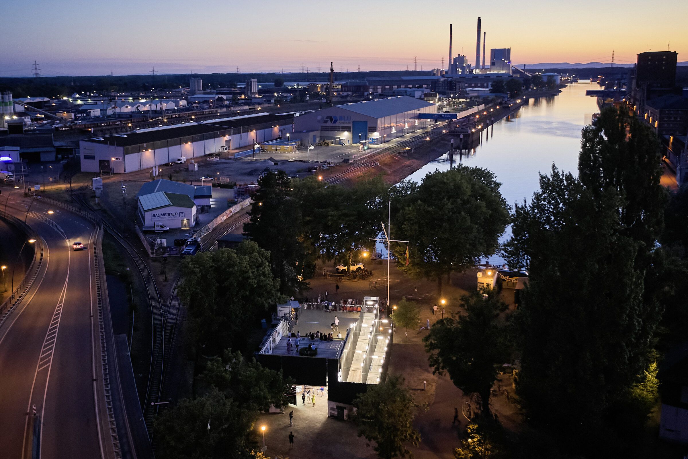 Luftbild in der Dämmerung: Der beleuchtete Bau im Vordergrund, dahinter das Hafenbecken und die Industrieanlagen des Rheinhafens bis zum Horizont.