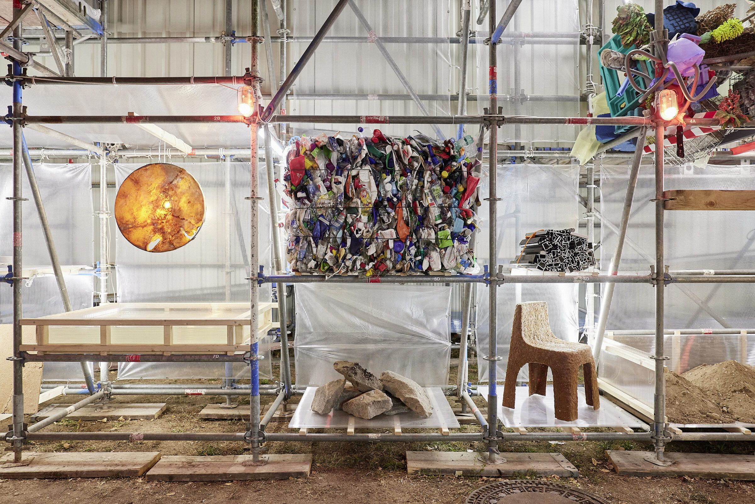 Das Gerüstregal der Materialbibliothek bei Nacht mit gepressten Kunststoffballen, einer orangefarbenen Scheibe und Materialproben.