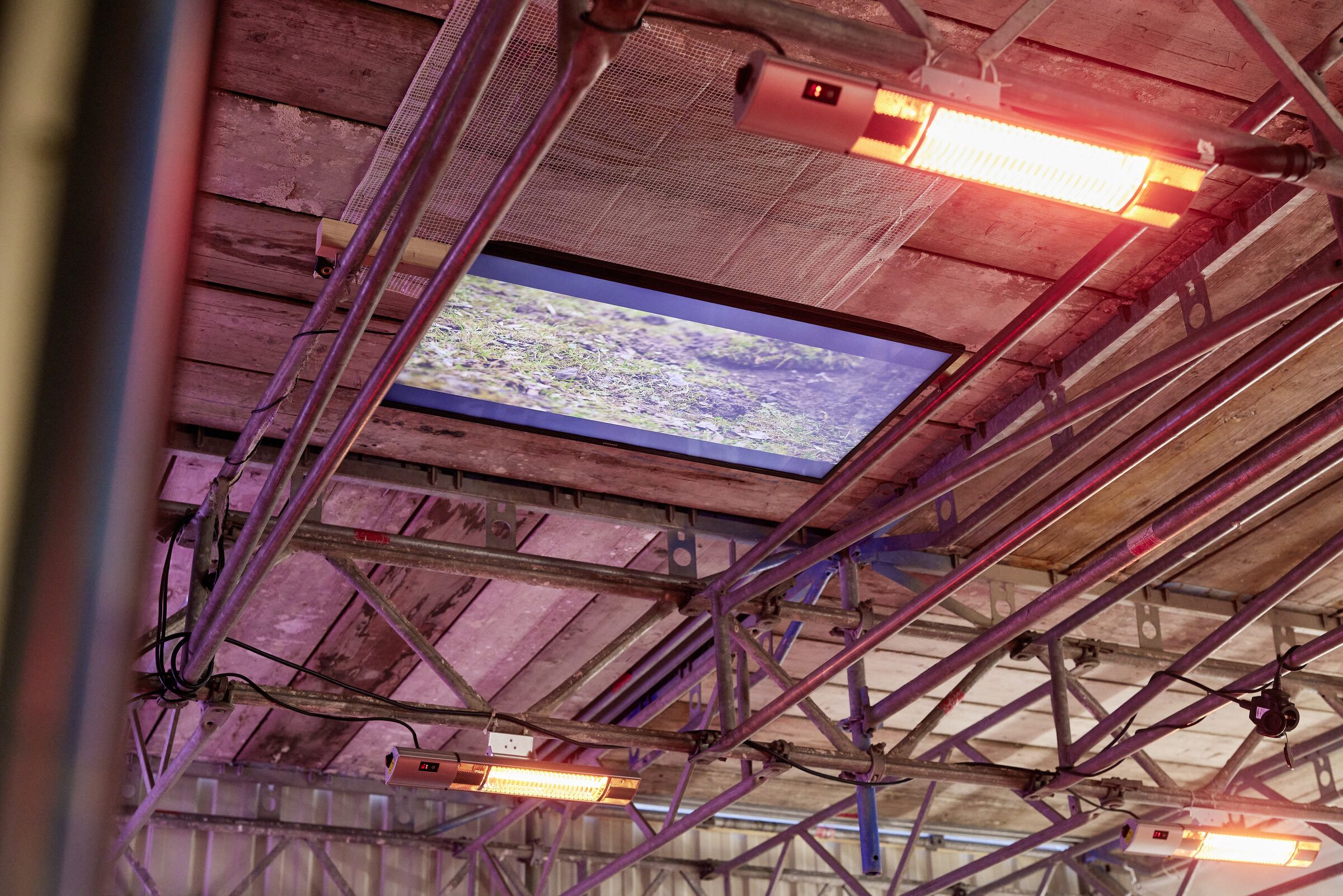 Ein Bildschirm hängt unter der Gerüstdecke zwischen roten Heizstrahlern.