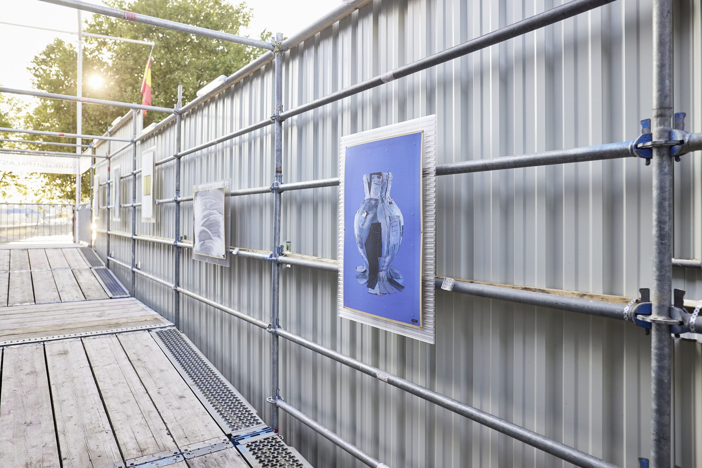 Plakate der Print-Edition hängen an der Gerüstwand entlang eines Holzstegs.